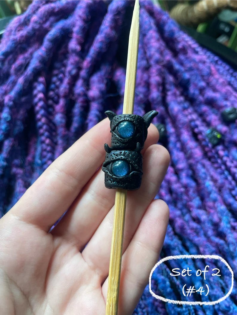 May include: A pair of black, horned dreadlock beads with blue, glittery eye details are displayed on a wooden stick. The beads are stacked, and the background features purple and blue dreadlocks. The image includes the text "Set of 2 (#4)".