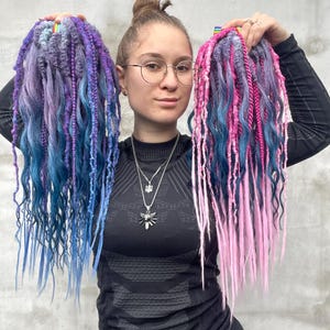 Puede incluir: Dos conjuntos de rastas coloridas en tonos azules, morados y rosas. Las rastas se sostienen en alto, mostrando las vibrantes combinaciones de colores y texturas. Las extensiones de cabello parecen hechas a mano.