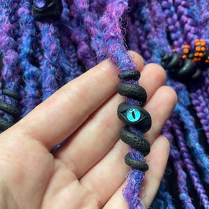 May include: Close-up of purple and blue dreadlocks with black decorative beads. One bead is a snake with a blue eye. The dreadlocks are a mix of purple, blue, and pink fibers.