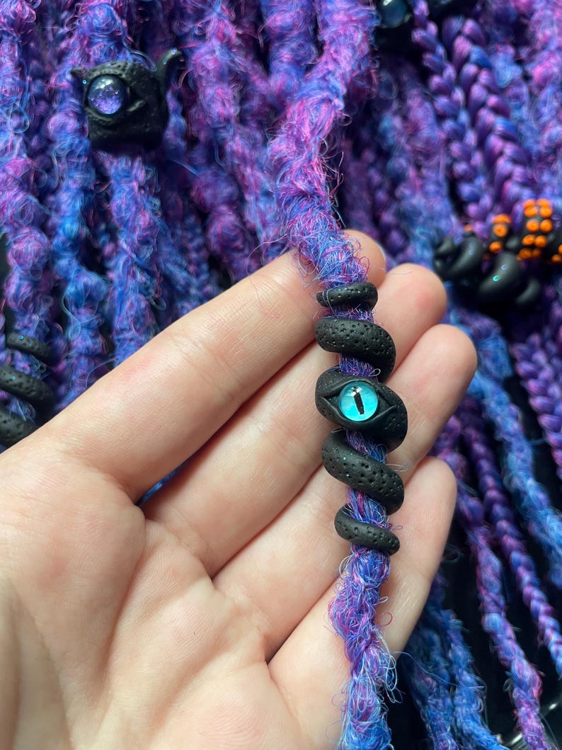 May include: Close-up of purple and blue dreadlocks with black decorative beads. One bead is a snake with a blue eye. The dreadlocks are a mix of purple, blue, and pink fibers.