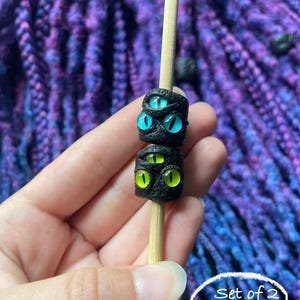 May include: Two black, textured beads with glowing eye designs, one with blue eyes and the other with green eyes, are displayed on a wooden skewer. The text "Set of 2 (#5)" is in a white circle.