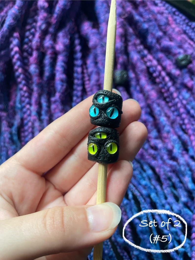 May include: Two black, textured beads with glowing eye designs, one with blue eyes and the other with green eyes, are displayed on a wooden skewer. The text "Set of 2 (#5)" is in a white circle.
