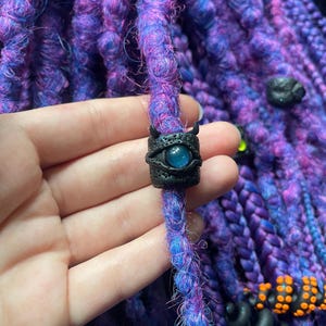 May include: Close-up of purple and blue dreadlocks with a black bead featuring a blue eye design. The bead has small horns and is held by a hand. Other black and orange beads are also visible.