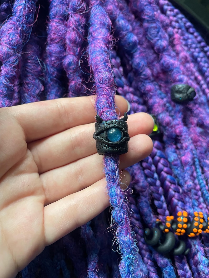 May include: Close-up of purple and blue dreadlocks with a black bead featuring a blue eye design. The bead has small horns and is held by a hand. Other black and orange beads are also visible.