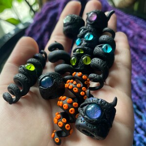 May include: A collection of black polymer clay sculptures with glowing, iridescent eyes in various colors, including blue, green, and purple. Some pieces feature orange tentacle-like details. The sculptures have a textured, porous surface.