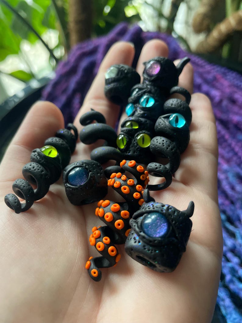 May include: A collection of black polymer clay sculptures with glowing, iridescent eyes in various colors, including blue, green, and purple. Some pieces feature orange tentacle-like details. The sculptures have a textured, porous surface.