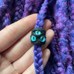 May include: A close-up of a purple and blue braided hair extension with a black bead featuring three blue eyes. The hair extension is a vibrant mix of purple and blue strands, creating a textured appearance. The bead is square-shaped.