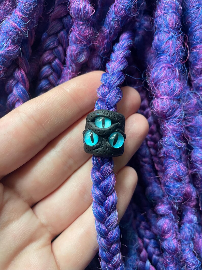 May include: A close-up of a purple and blue braided hair extension with a black bead featuring three blue eyes. The hair extension is a vibrant mix of purple and blue strands, creating a textured appearance. The bead is square-shaped.