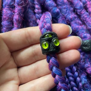 May include: A close-up of a braided purple and blue hair accessory with a black bead featuring three glowing green eyes. The background is a textured, purple and blue knitted fabric.