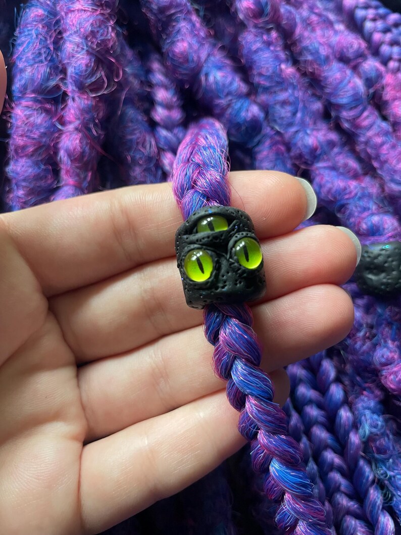 May include: A close-up of a braided purple and blue hair accessory with a black bead featuring three glowing green eyes. The background is a textured, purple and blue knitted fabric.