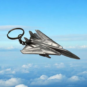 Puede incluir: Un llavero de metal plateado con forma de avión de combate. El avión está volando contra un cielo azul con nubes blancas.