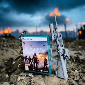 Peut inclure: Un porte-clés avec un jeu PlayStation 5 et un fusil miniature gris. Le jeu porte le titre "Battlefield 6" avec un graphisme de soldats. L'arrière-plan montre un ciel flou et dramatique.