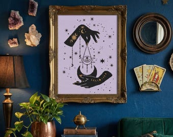 May include: A framed print with a gold ornate frame features black hands holding a crescent moon and celestial symbols on a lavender background. The print is displayed on a dark blue wall with other decorative items.
