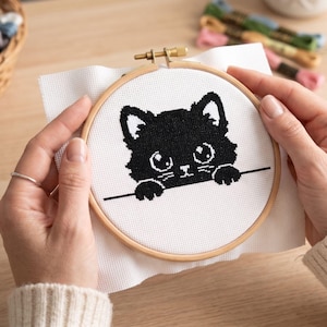 Op de afbeelding: Een zwart katten kruissteekontwerp in een houten borduurring. De kat heeft grote ogen en kijkt over een lijn. De achtergrond is witte stof. Borduurgaren in verschillende kleuren is zichtbaar op de achtergrond.