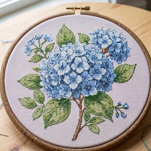 May include: A completed cross-stitch embroidery of a blue hydrangea flower with green leaves and a brown stem. The embroidery is set in a wooden hoop, showcasing the detailed needlework on a light pink fabric.