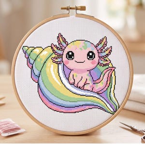 May include: A cross-stitch embroidery of a cartoon axolotl sitting in a rainbow-colored seashell. The axolotl is pink with colorful accents, and the seashell features pastel hues. The embroidery is framed in a wooden hoop.