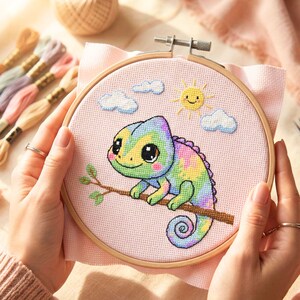 May include: A cross-stitch embroidery hoop featuring a colorful chameleon perched on a branch. The design includes a smiling sun and fluffy clouds against a pink background. The wooden hoop is held by hands.
