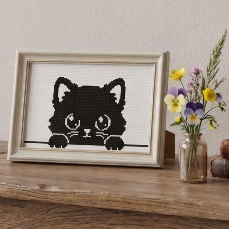 May include: A framed cross-stitch art piece featuring a black cat silhouette with large eyes and paws peeking over a line. The artwork is in a white frame, displayed on a wooden surface, with a small vase of flowers nearby.