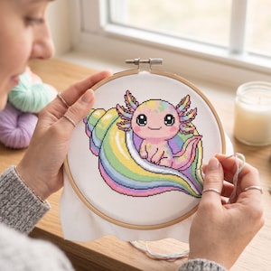 May include: A cross-stitch project featuring a colorful axolotl sitting in a rainbow seashell. The axolotl is pink, yellow, and blue. The project is in a wooden embroidery hoop, with the needle and thread visible.