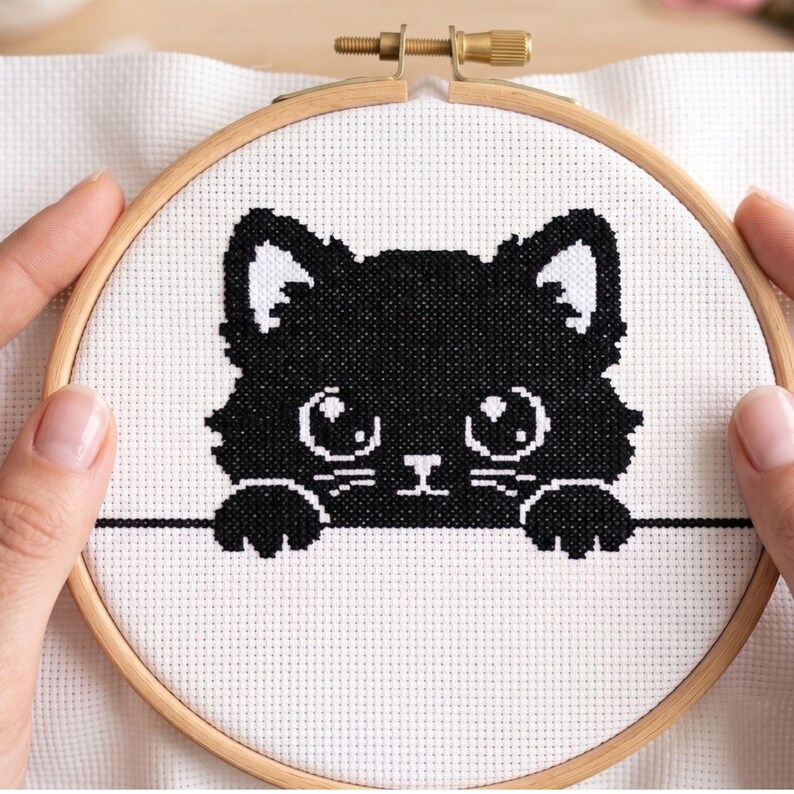 May include: A cross-stitch project featuring a black cat design. The cat has large eyes and is framed by a wooden embroidery hoop. The cat's silhouette is stitched on white fabric. The hoop is held by hands.