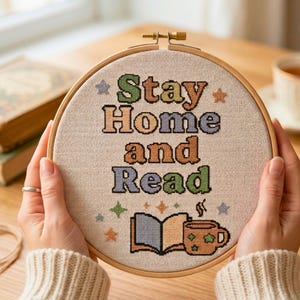 May include: A cross-stitch embroidery hoop with the words "Stay Home and Read" in colourful letters. The design includes an open book and a mug. The hoop is held by hands wearing a ring and a cream-coloured sweater.