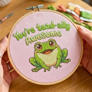 May include: Cross-stitch embroidery hoop featuring a green frog with the text "You're toad-ally Awesome" in green. The frog has large brown eyes and a smiling expression. The background is light pink, and the hoop is made of wood.