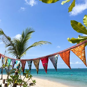 May include: A string of colorful triangular flags hangs against a backdrop of a tropical beach. The flags, in shades of red, orange, blue, and green, are strung along a rope. The beach has white sand, turquoise water, and a clear blue sky.