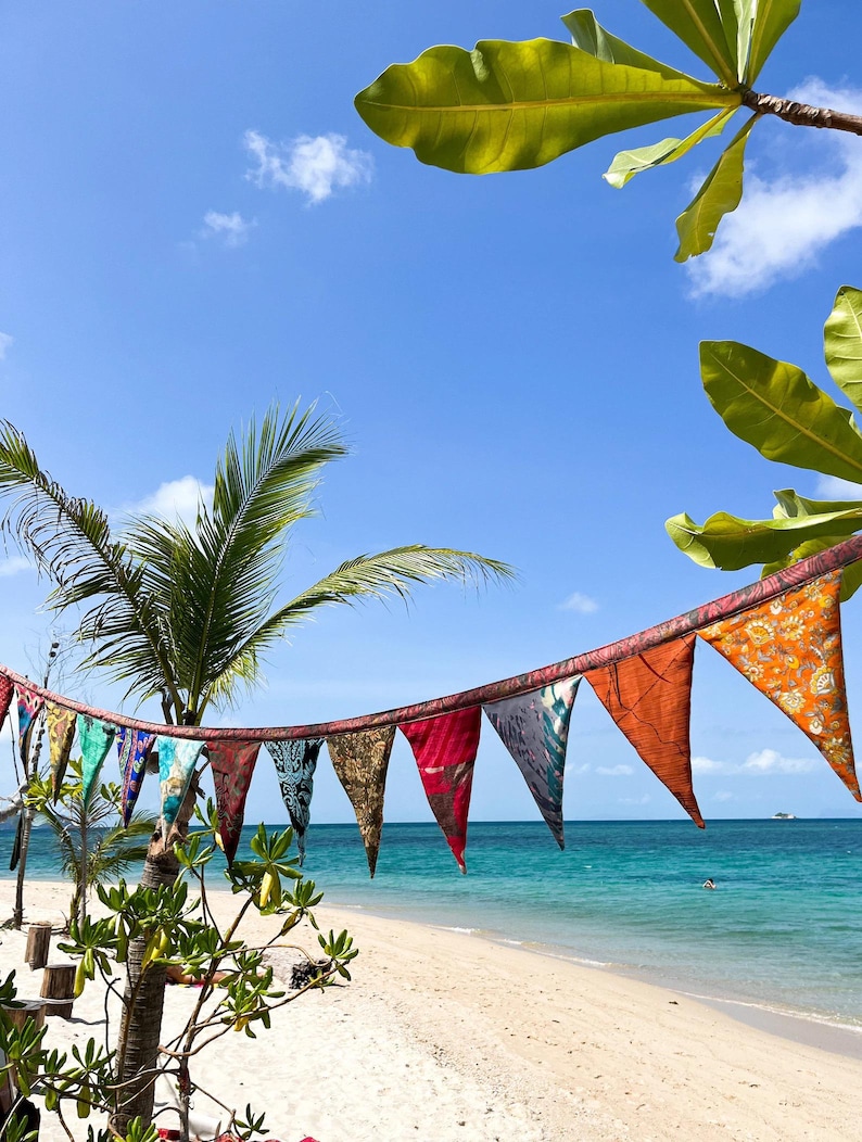 May include: A string of colorful triangular flags hangs against a backdrop of a tropical beach. The flags, in shades of red, orange, blue, and green, are strung along a rope. The beach has white sand, turquoise water, and a clear blue sky.