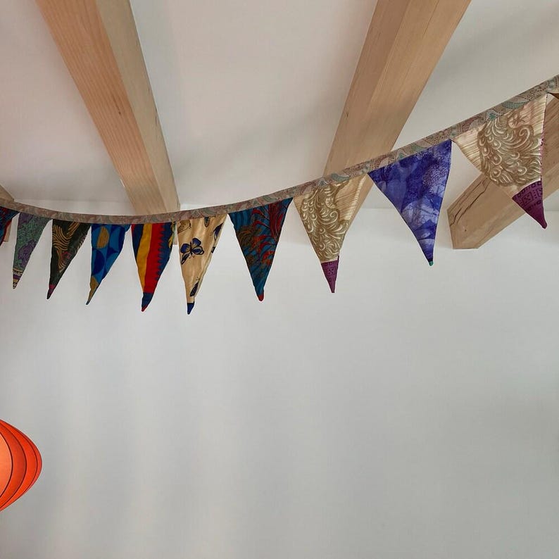 May include: A colorful fabric banner with triangular pennants. The pennants feature various patterns and colors, including blue, gold, red, and purple. The banner is strung across a room with wooden beams and a white wall.