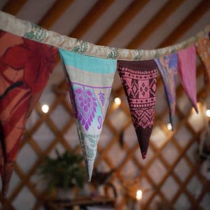 May include: A colorful fabric banner with triangular pennants hangs from a ceiling. The pennants feature various patterns and colors, including red, teal, pink, and purple. The background shows a wooden structure and soft lighting.