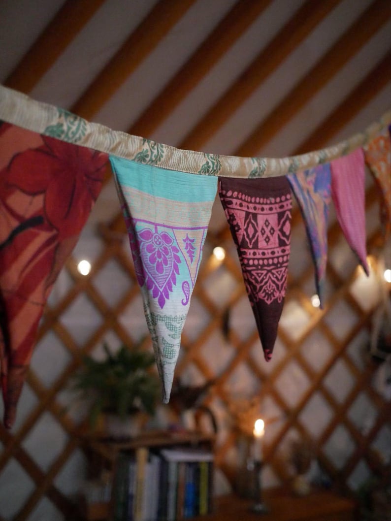May include: A colorful fabric banner with triangular pennants hangs from a ceiling. The pennants feature various patterns and colors, including red, teal, pink, and purple. The background shows a wooden structure and soft lighting.