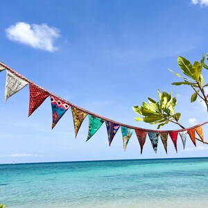 May include: A string of colorful fabric pennant flags hangs against a bright blue sky and turquoise ocean. The flags are triangular, with various patterns and colors, including red, green, and blue. A small tree with green leaves is on the right.