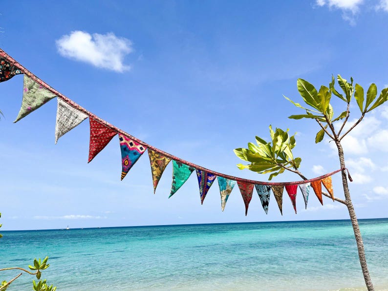 May include: A string of colorful fabric pennant flags hangs against a bright blue sky and turquoise ocean. The flags are triangular, with various patterns and colors, including red, green, and blue. A small tree with green leaves is on the right.