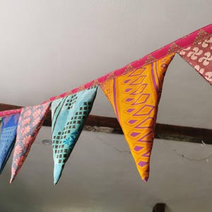 May include: A colorful fabric bunting with triangular flags. The flags display various patterns and colors, including blue, pink, yellow, and brown. The bunting is strung along a pink ribbon, creating a festive and decorative element.