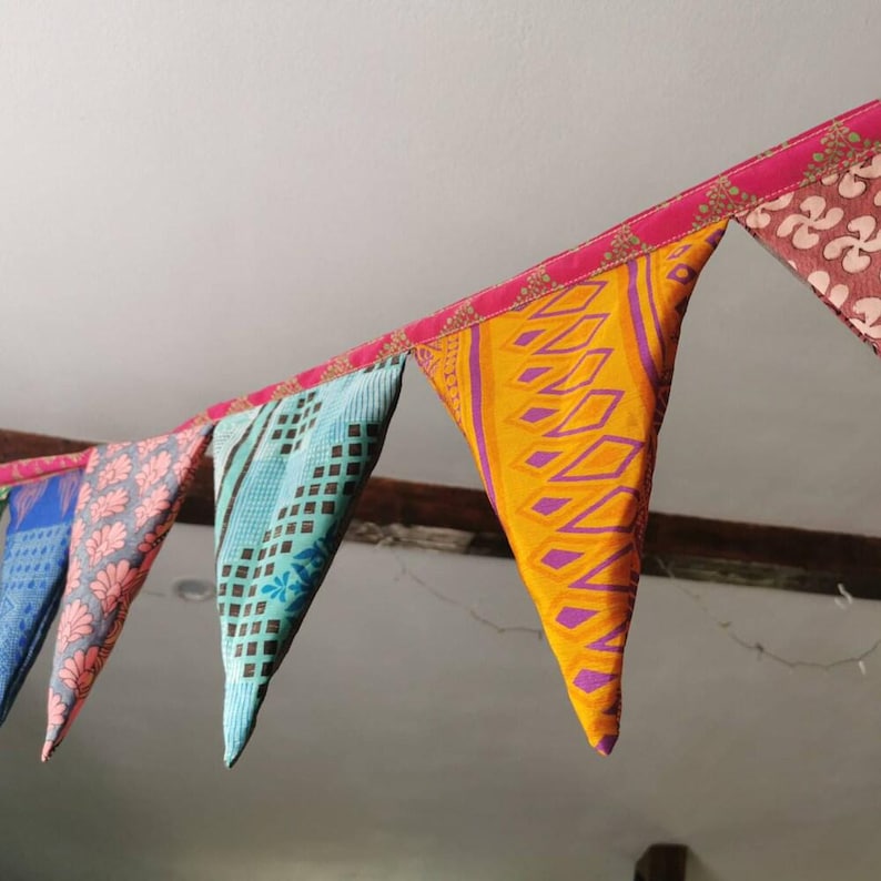 May include: A colorful fabric bunting with triangular flags. The flags display various patterns and colors, including blue, pink, yellow, and brown. The bunting is strung along a pink ribbon, creating a festive and decorative element.