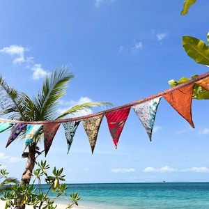 May include: A string of colorful triangular flags hangs against a bright blue sky with fluffy white clouds. The flags are various colors and patterns, with a beach and ocean in the background.