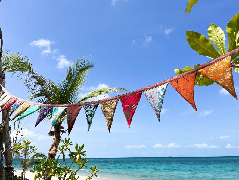 May include: A string of colorful triangular flags hangs against a bright blue sky with fluffy white clouds. The flags are various colors and patterns, with a beach and ocean in the background.