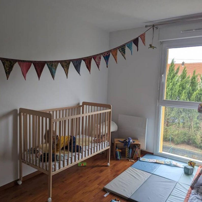 May include: A light wood baby crib with a child inside, set in a room with a wooden floor. A colorful pennant banner hangs above the crib. The room has a window with a view of trees and a blue and gray play mat.