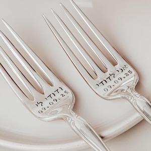 May include: Two silver forks with ornate handles, resting on a white plate. Each fork is engraved with text and the date 09.09.25. The forks are placed on a white plate with a rust-colored cloth in the background.
