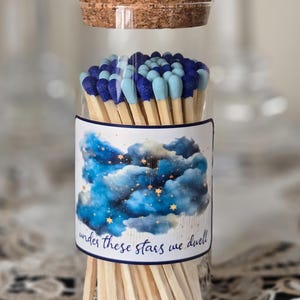May include: A clear glass jar with a cork stopper filled with wooden matches that have dark blue and light blue tips. A label on the jar features a blue watercolor cloud and star illustration with the text "under these stars we dwell".