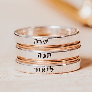 May include: A stack of three silver and rose gold rings. Each ring has a band of silver with a rose gold band in the middle. The rings have Hebrew text engraved on them. The rings are stacked on top of each other.