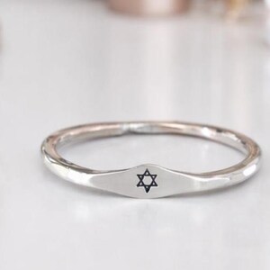 May include: A silver ring featuring a Star of David. The ring has a smooth, rounded band with a flat, rectangular face bearing a black Star of David engraving. The ring is shown on a white surface.