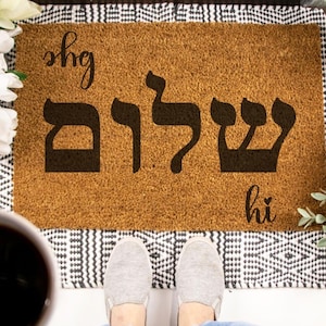 May include: A rectangular doormat with the Hebrew word "Shalom" in large black letters. The words "bye" and "hi" are written in cursive on the mat. The mat is surrounded by a black and white patterned border.