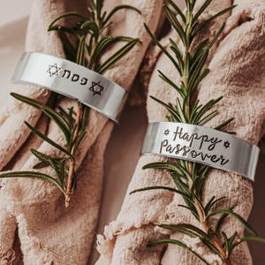 May include: Two silver napkin rings with text and star designs. One ring says "Happy Passover" and the other has Hebrew text. The rings are wrapped around beige napkins and decorated with sprigs of rosemary.