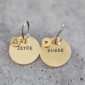 May include: Two gold keychains with round metal tags. The tag on the left says "ZEYDE" and has a small heart charm with the letter "B" on it. The tag on the right says "BUBBE" and has a small round charm with the letter "A" on it.