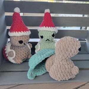 May include: Three crocheted stuffed toys wearing Santa hats. The toys are a brown turtle, a green turtle, and a brown turtle with a green shell.