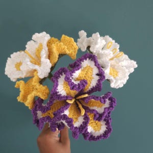 Puede incluir: Un ramo de flores de ganchillo hechas a mano en blanco, amarillo y morado. Las flores tienen diseños de pétalos detallados y se sostienen sobre un fondo turquesa. El trabajo de ganchillo es intrincado, mostrando la textura y la artesanía.