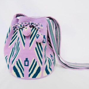 Op de afbeelding: Een lavendelkleurige gehaakte tas met een geometrisch patroon in wit en turquoise. De tas heeft een lange band en een trekkoord met kwastjes.
