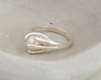 May include: A silver ring with a white pearl set in a unique, abstract design. The ring is made of smooth, polished metal and features a delicate, intricate design.