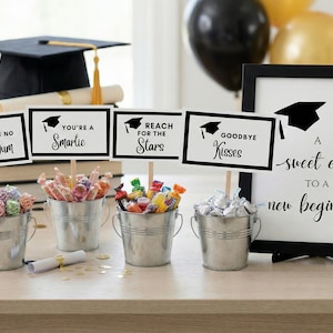 Puede incluir: Decoraciones para fiestas de graduación. Incluye un birrete negro, letreros con frases como "You're a Smartie" y "Goodbye Kisses", y pequeños cubos de metal llenos de caramelos. Un letrero enmarcado dice "A sweet ending to a new beginning!"